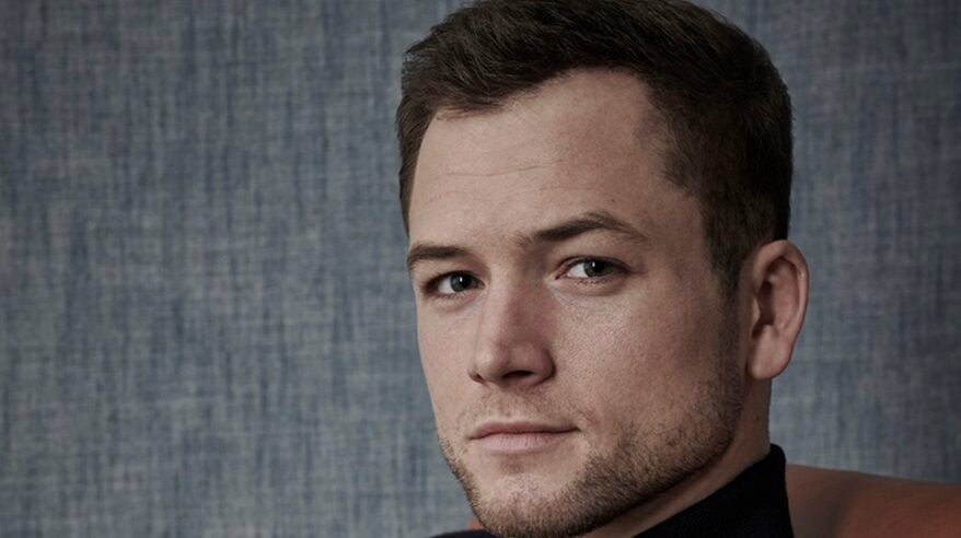 Actor Taron Egerton looks sideways to camera, with a grey background