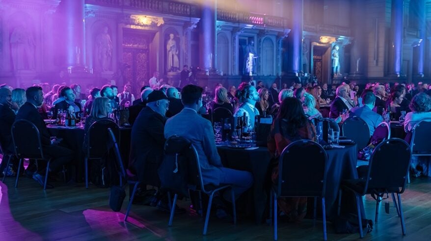 A dark and elegantly lit room with formal round tables and people smartly dressed for evening event