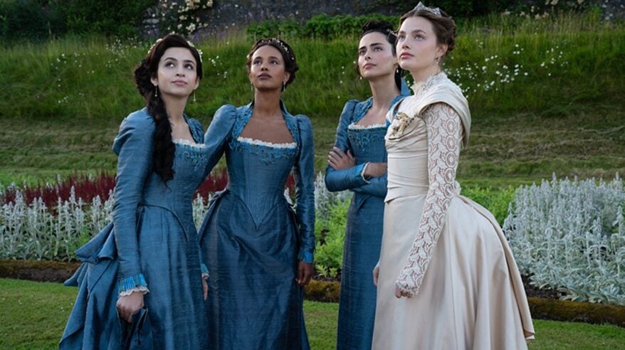 Four ladies in a group outdoors, with foliage in the background, look up to the sky, wearing elegant late 19th century dresses.