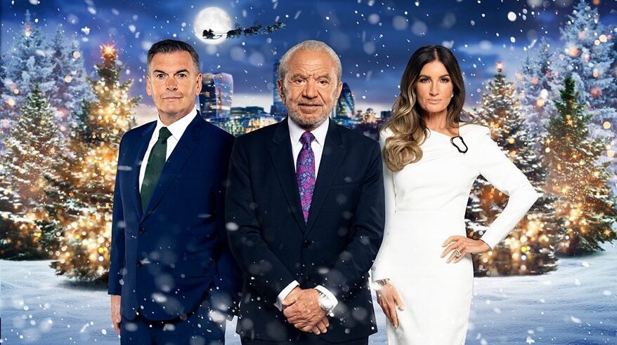 Two smartly-dressed men and a women in a glamorous white dress stand in front of a snowy, nighttime Christmas background with reindeers and sleigh silhouetted behind