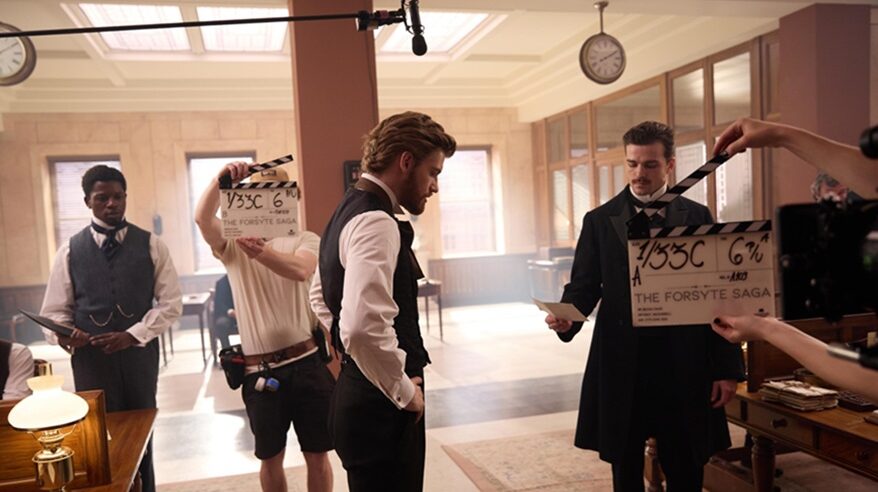A man in 19C clothing stands in a light room, with camera crew holding kit and clapperboards around him