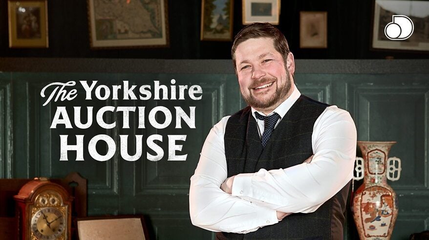 Bearded man in shirt, tie and waistcoat stands with arms folded, smiling, with the words The Yorkshire Auction House behind him