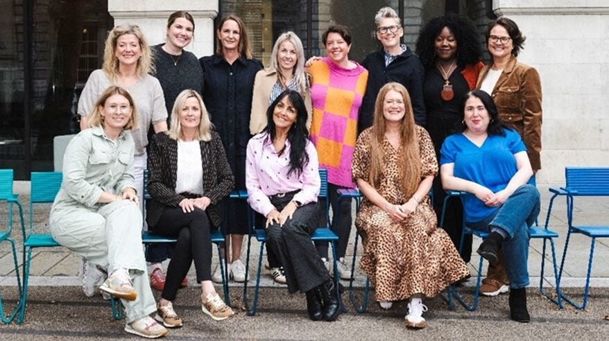 Group of 12 women with the front row seated in blue chairs, outdoors with stone pillars in background
