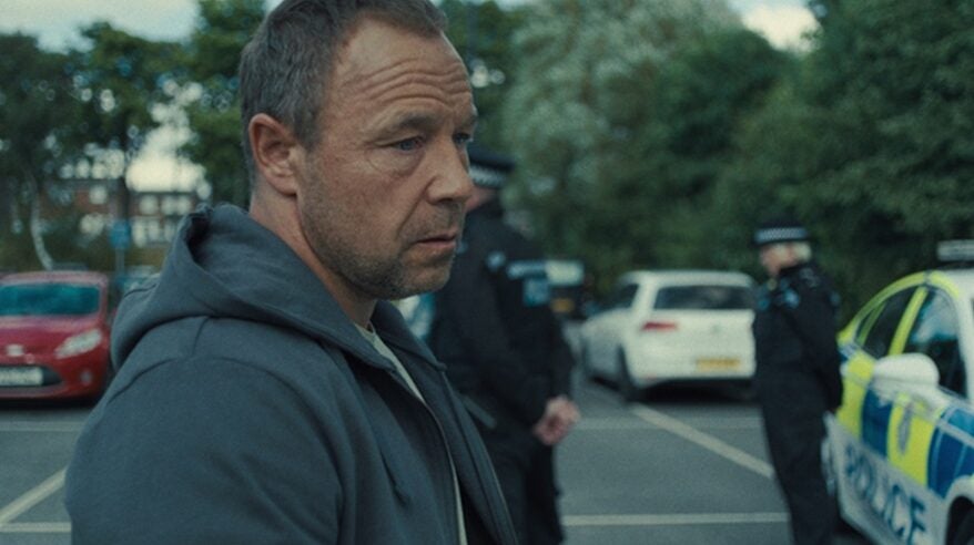 A man stands in a grey hoodie, looking sadly at the ground of a car park with police presence behind him