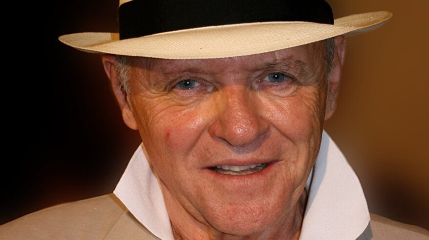 Blue-eyed man in straw fedora and white shirt collar smiles to camera
