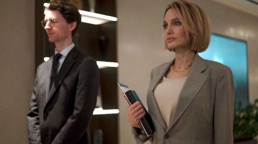 A smartly-dressed woman in a pale business jacket with a blonde bob stands in an office next to a man in a dark suit, from the film Anxious People, shot at Pinewood's Indie Film Hub