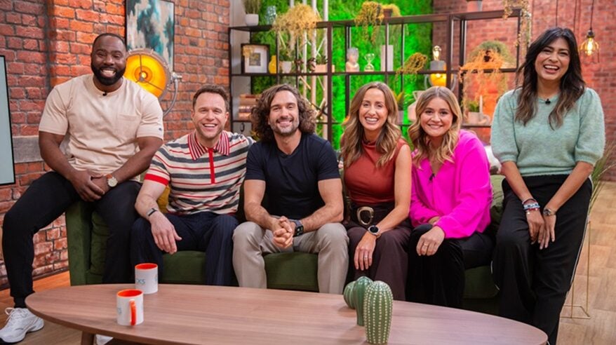 A group of smiling people sit together on a sofa, with a bookshelf and bright green wall behind and a coffee table in front