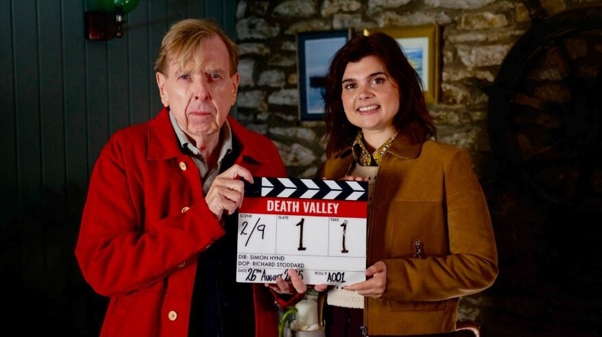 A fair-haired man in a red jacket and a dark-haired lady in a brown jacket jointly hold a film clapperboard