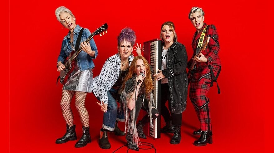 Five women dressed as punk rockers hold instruments and mikes in front of an orange background