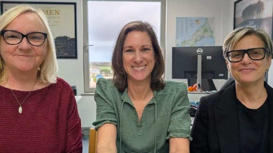 Two blonde women sit either side of a dark-haired lady at a desk in an office with maps on the wall behind
