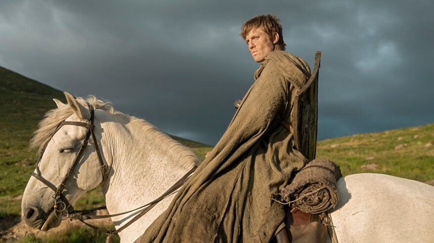 A tall man on a white horse in medieval-looking brown sackcloth robe looks outwards on a hill with grey skies behind