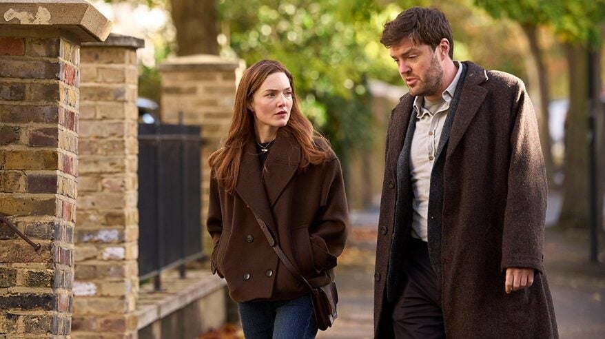 A woman with long red hair in a brown jacket walks down a street next to a dark haired, bearded man in a brown coat