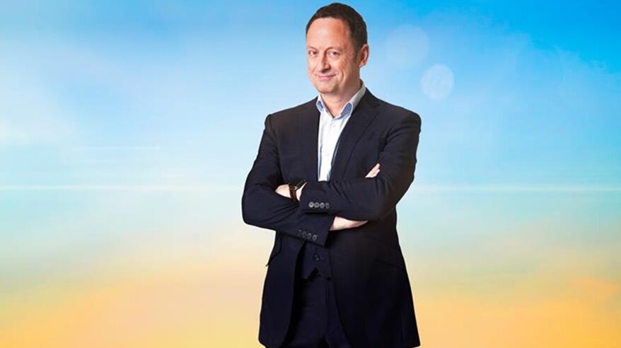 A dark-haired man in a black suit with white open-necked shirt stands smiling with his arms folded, against a blue and yellow ombre wall