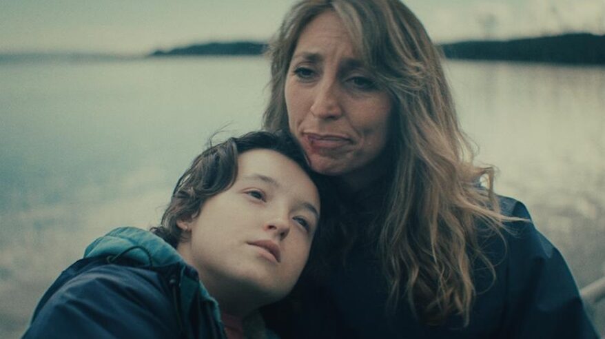 Two people sit close together in front of a loch, one teenage and one lady with long hair, in a melancholy shot with grey overtones