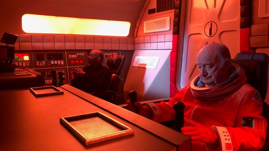 Two men sit in the cockpit of a model of a spaceship, the nearer one in a spacesuit